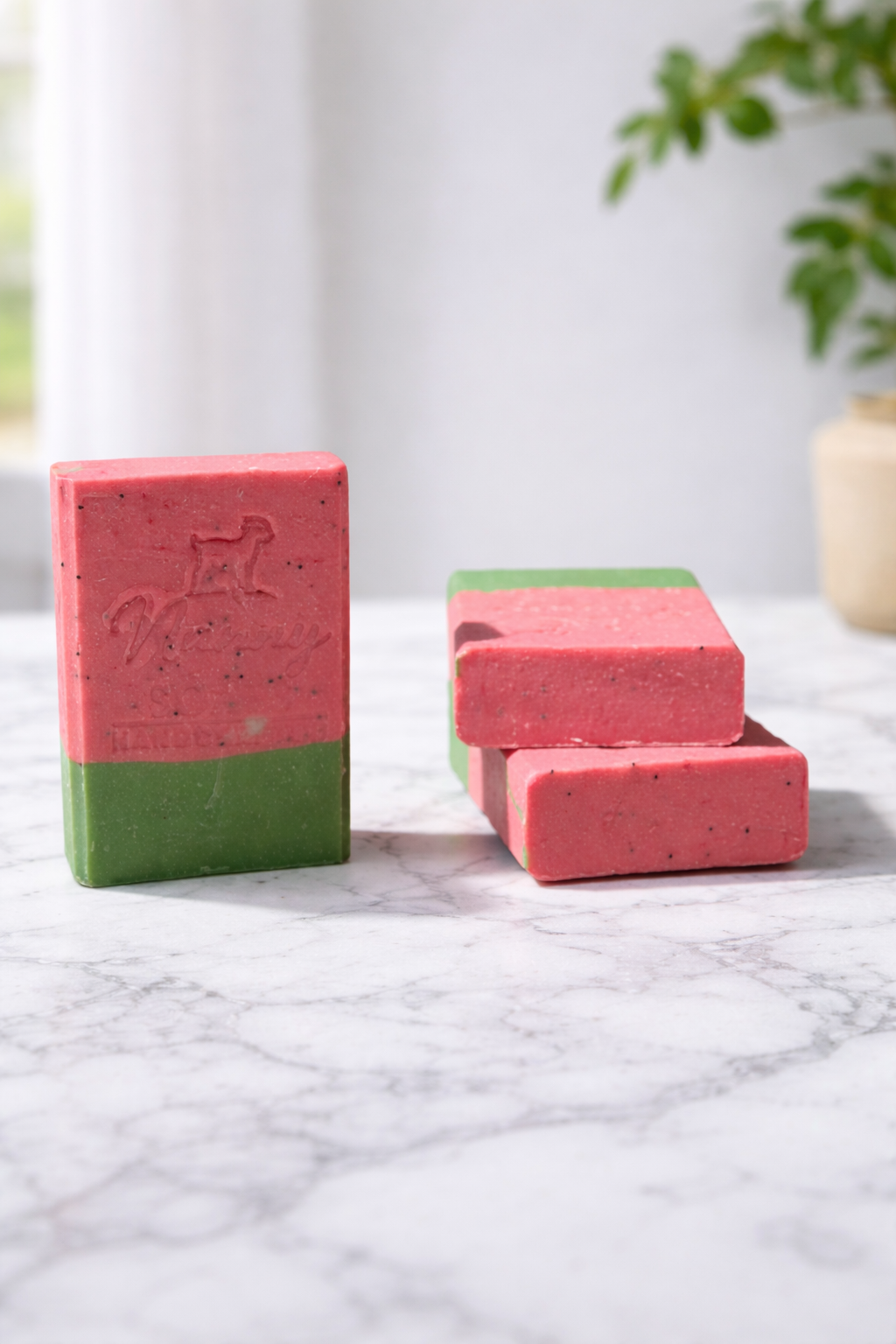 Two pink and green soap bars on a marble surface with a blurred plant in the background