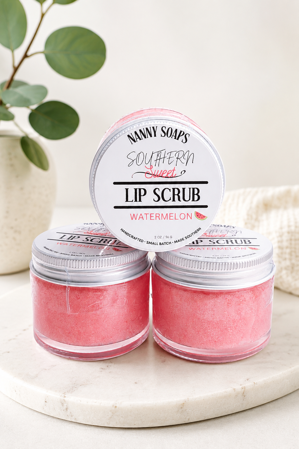 Three small jars of pink lip scrub with silver lids are arranged on a round marble tray. One jar is open and placed behind the two closed jars, showing the label 'Nanny Soap's Southern Sweet Lip Scrub Watermelon.